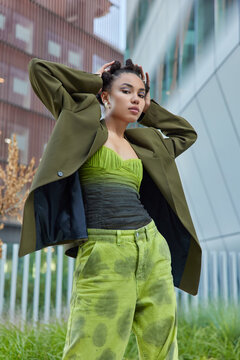 Outdoor shot of fashionable young woman stands in self confident pose keeps hands behind head dressed in stylish clothes looks seriously at camera stands against blurred city background. Street style