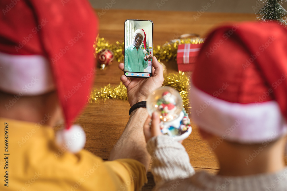 Father and son in santa hats making smartphone christmas video call with happy african american man