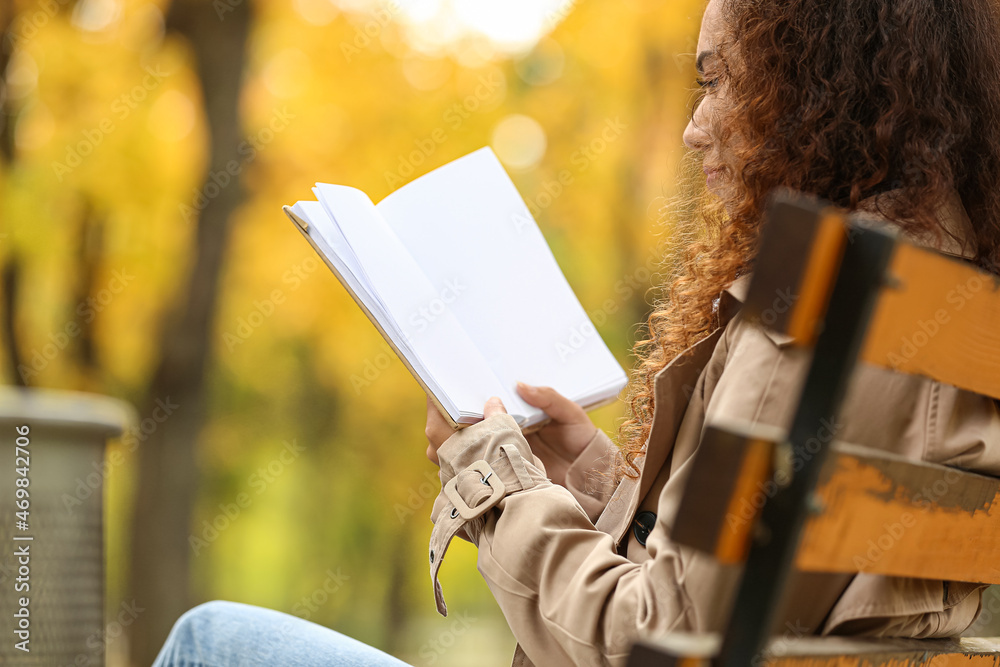 年轻的非裔美国妇女在秋季公园的长椅上阅读一本空白页的书