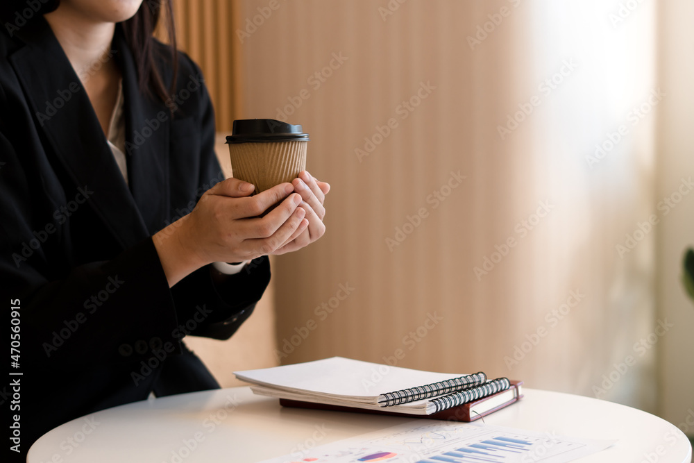 在靠近窗户的办公室工作期间，商界女性的手拿着一杯热咖啡。