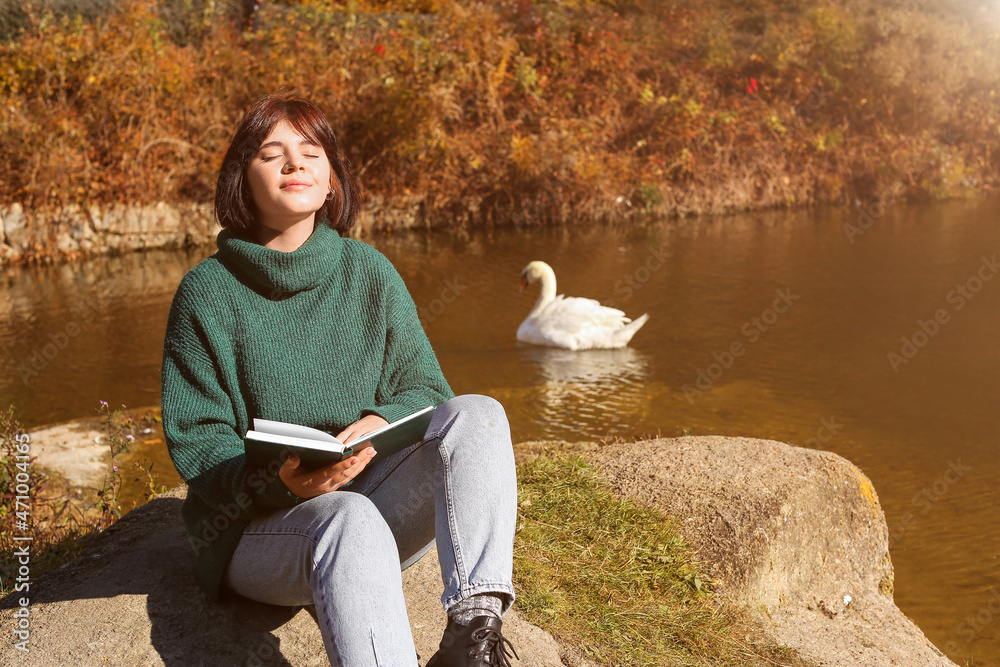 带着书的年轻女子与天鹅在池塘附近享受阳光明媚的一天