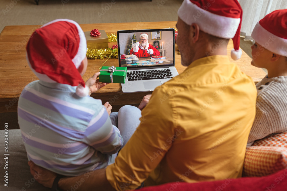 戴着圣诞老人帽的父亲和儿子与微笑的圣诞老人进行笔记本电脑圣诞视频通话