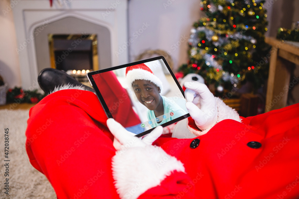 Santa claus making tablet christmas video call with smiling african american boy in santa hat