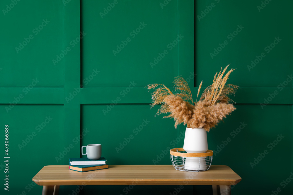 绿色墙壁附近的长椅上放着干芦苇、杯子和书的花瓶