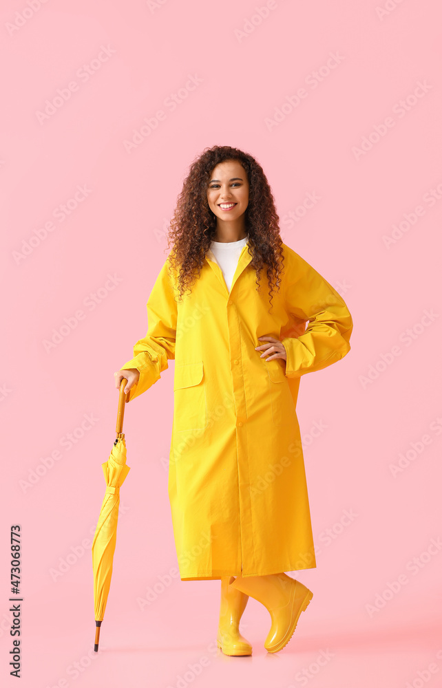 年轻的非裔美国妇女穿着黄色雨衣和胶靴，粉色背景下有伞