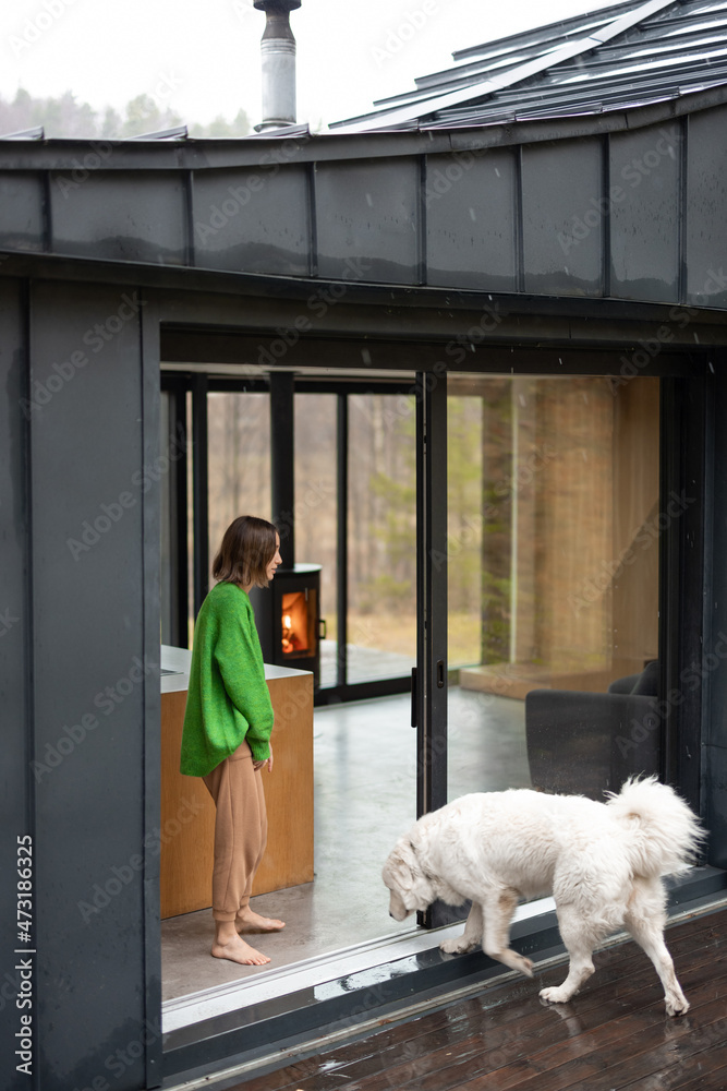 Young woman with her dog at modern house on nature. Girl in green sweater standing near the window a