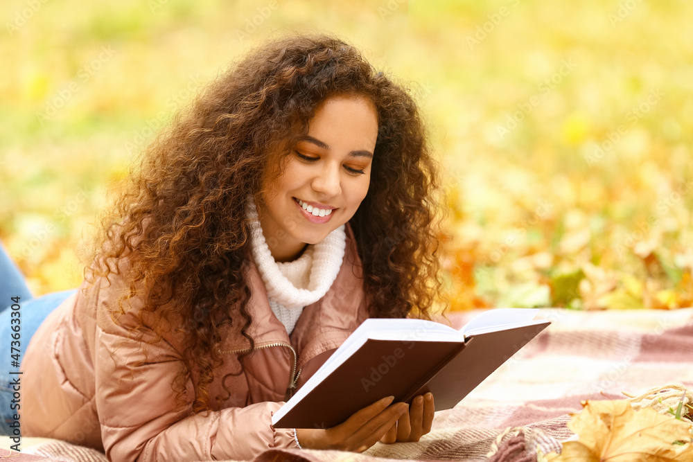 年轻微笑的非裔美国妇女在秋季公园读格子书
