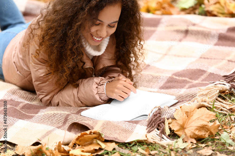 一位年轻的非裔美国妇女在秋天的公园里阅读一本格子空白页的书