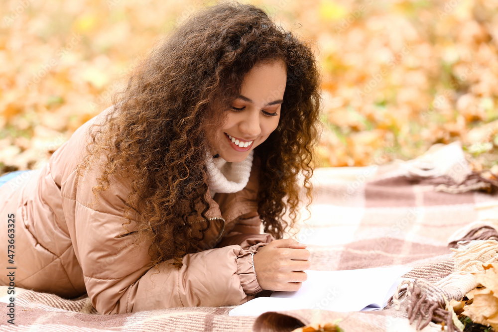 美丽的非裔美国女人在秋天的公园里阅读一本格子空白页的书