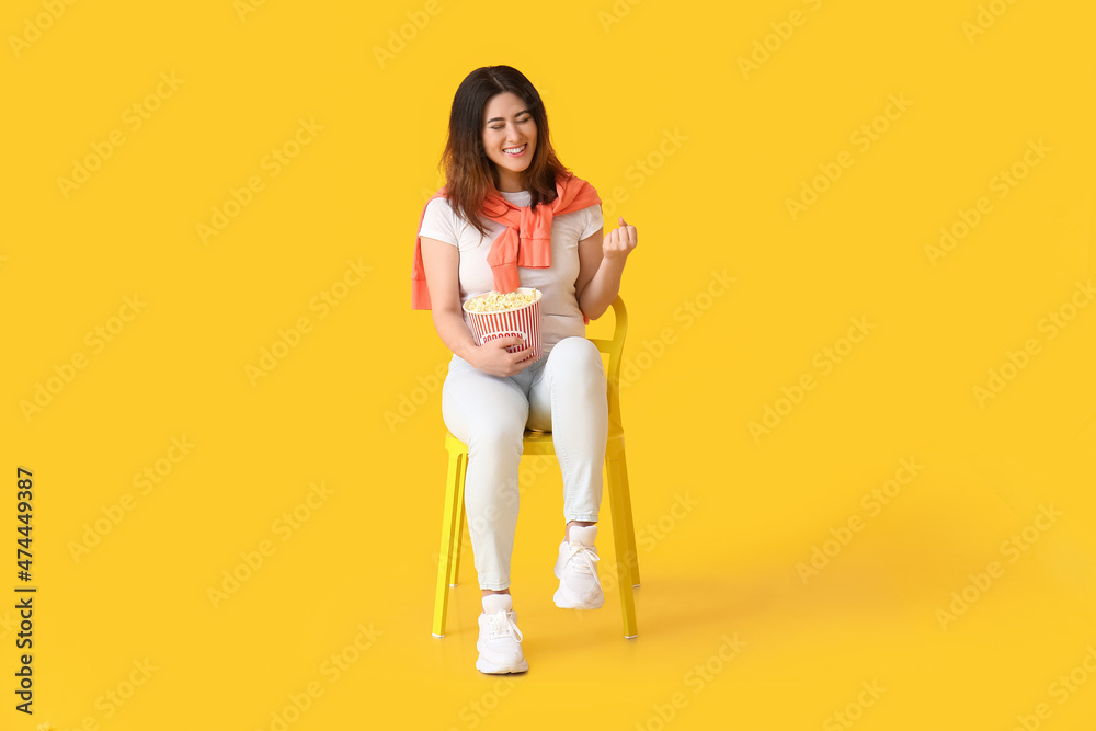 Happy young Asian woman with bucket of tasty popcorn sitting in chair on yellow background