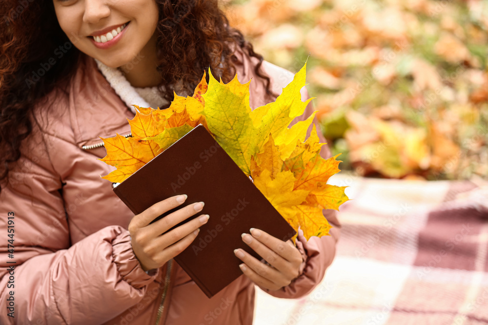 Beautiful African-American woman holding book with dry leaves in autumn park, closeup