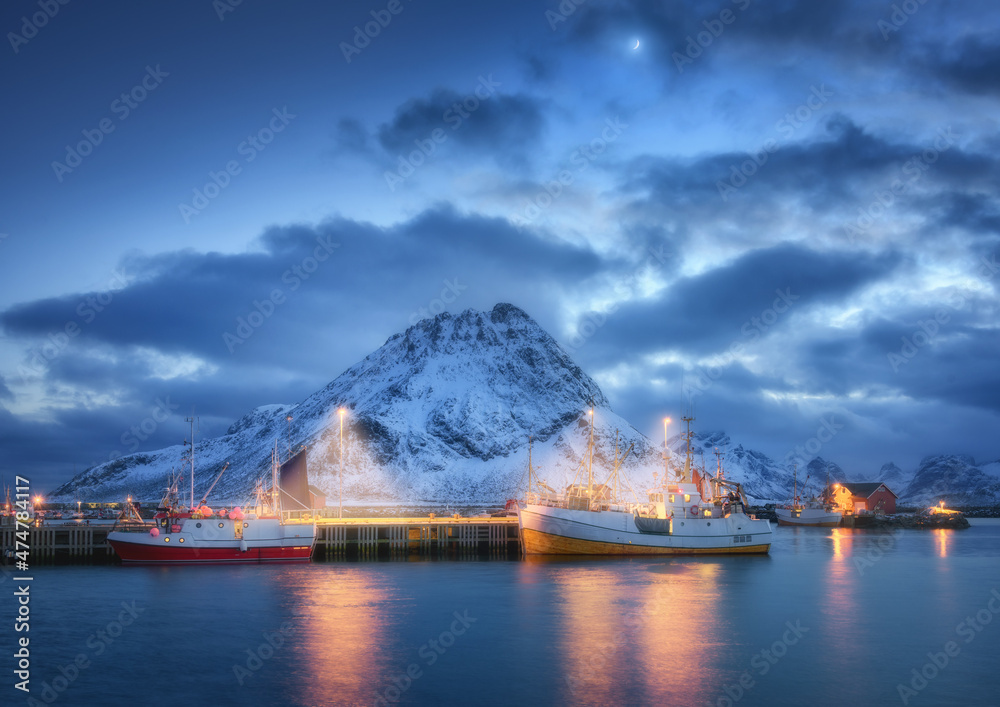 Fishing boats on the sea, snowy mountains, sky with clouds, moon at night in Lofoten islands, Norway