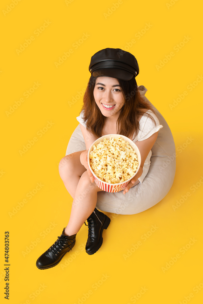年轻的亚洲女人拿着美味的爆米花坐在黄色背景的豆袋椅上