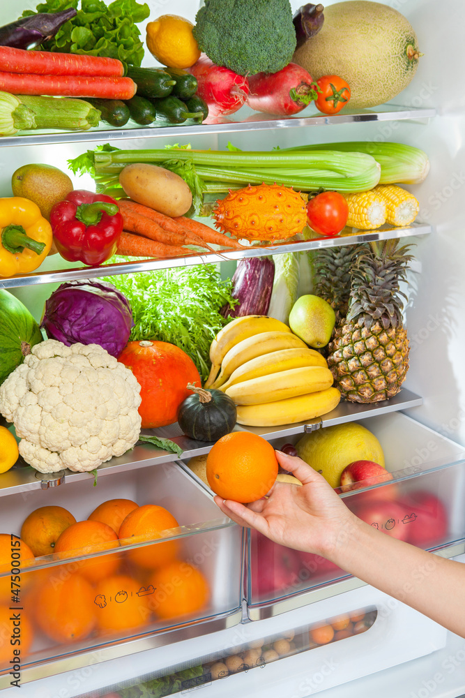 女人的手从装满水果和蔬菜的冰箱里拿出一个橙子。