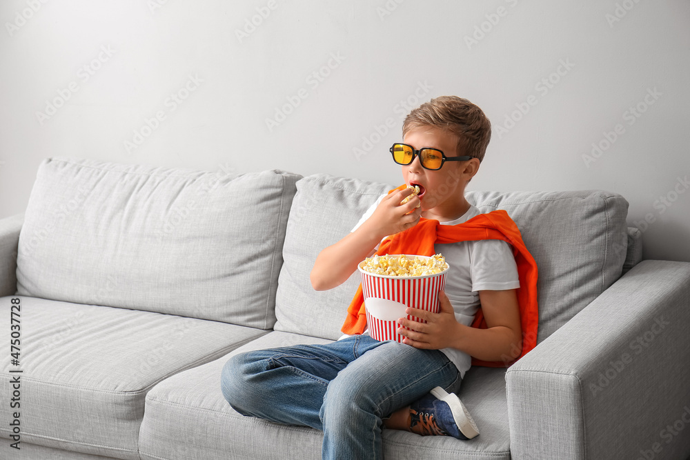 戴眼镜的小男孩在灯光墙附近的沙发上吃美味的爆米花