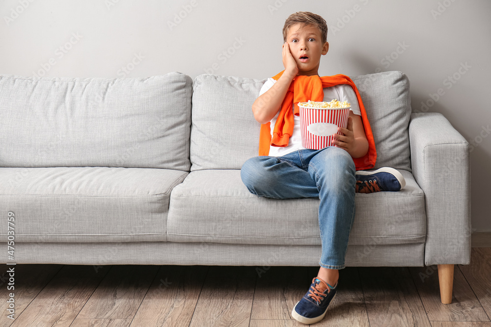 震惊的小男孩拿着一桶美味的爆米花坐在靠近灯光墙的沙发上
