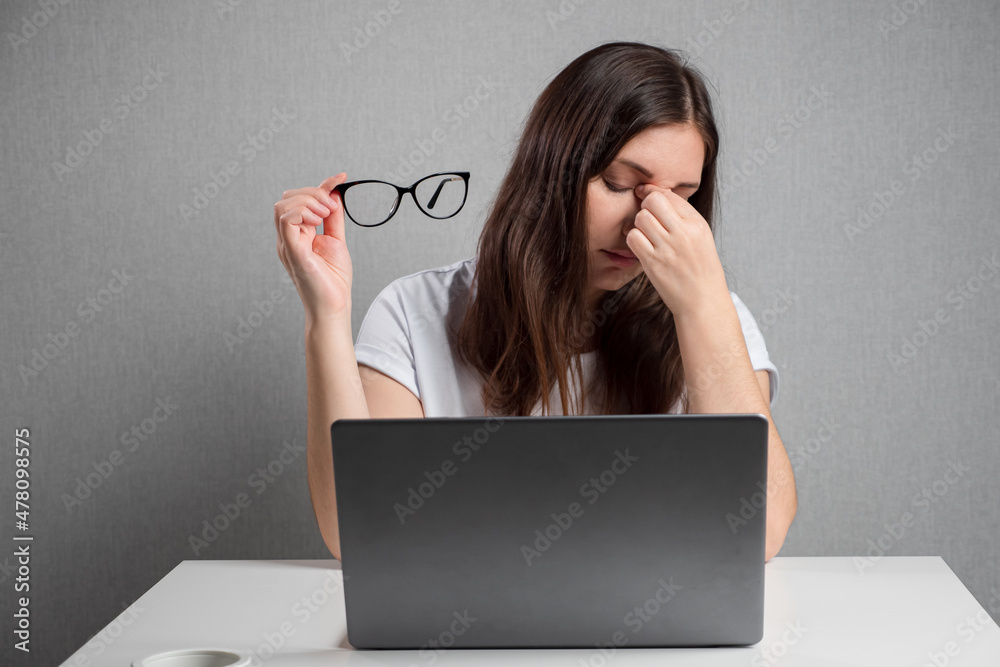 穿着白色t恤衫的疲惫女子摘下眼镜，坐在笔记本电脑屏幕前用手指摩擦酸痛的眼睛