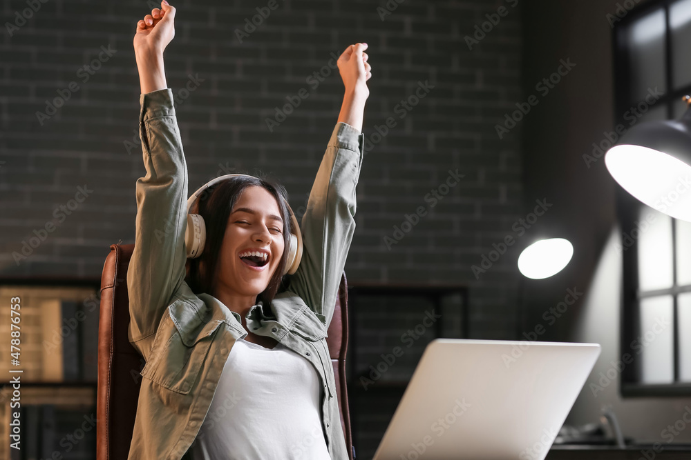 Happy young woman with laptop listening to music at home in evening