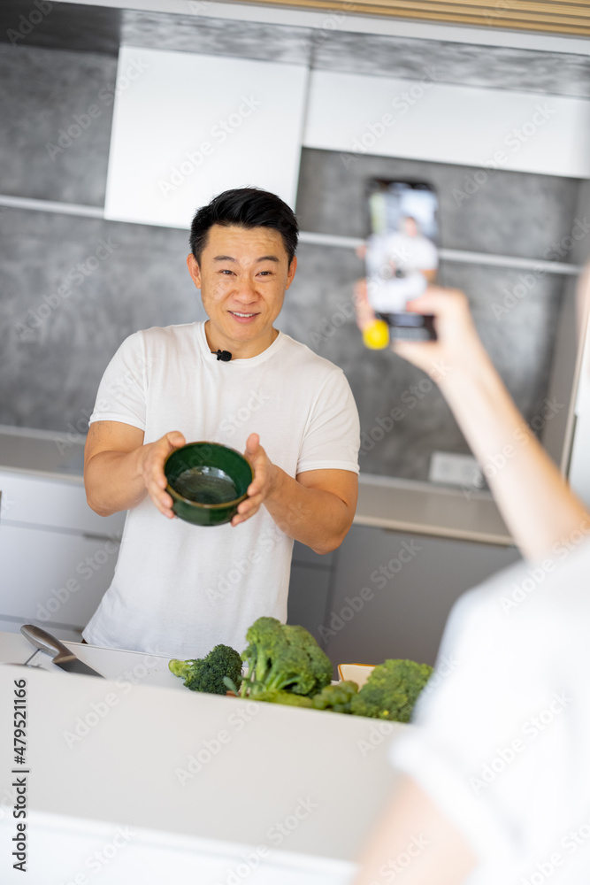 一名女子在智能手机上拍摄亚洲男子在厨房烹饪素食沙拉的视频。视频的概念