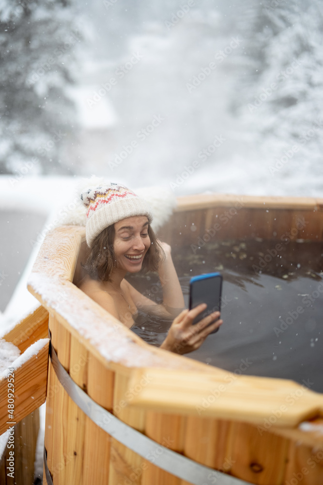 快乐的女人在室外洗热水澡放松，在下雪的时候坐在温泉里打电话