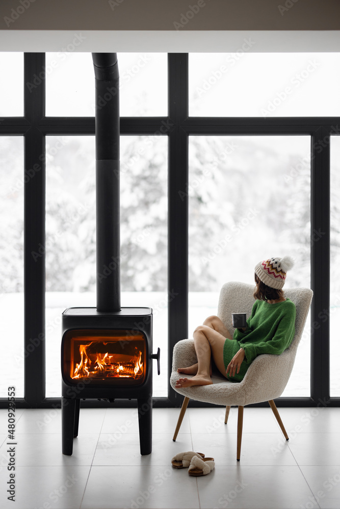 Woman sitting with cup on chair by the fireplace at modern house on nature during winter time. Conce