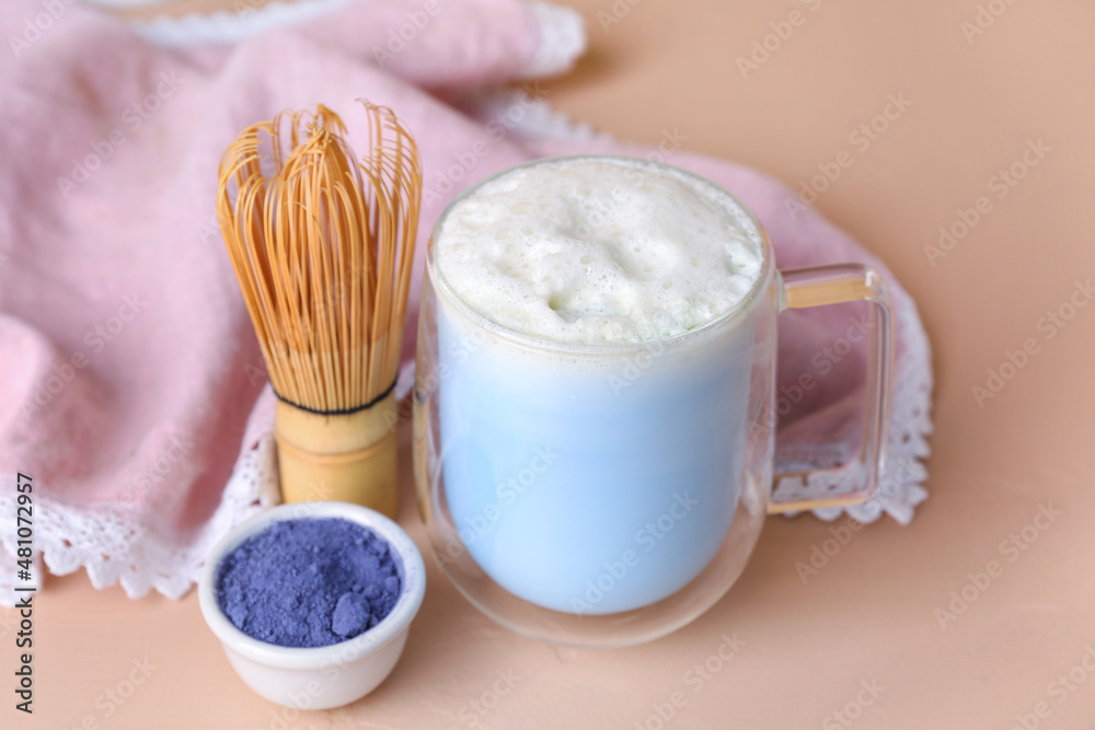 Glass cup of blue matcha latte, chasen and bowl with powder on color background