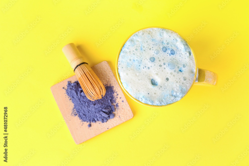 Cup of blue matcha latte, powder and chasen on yellow background