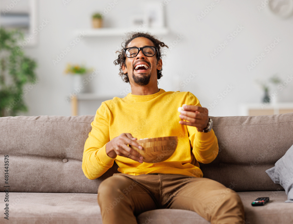 年轻的非裔美国人拿着爆米花大笑，在家看喜剧电影，欣喜若狂
