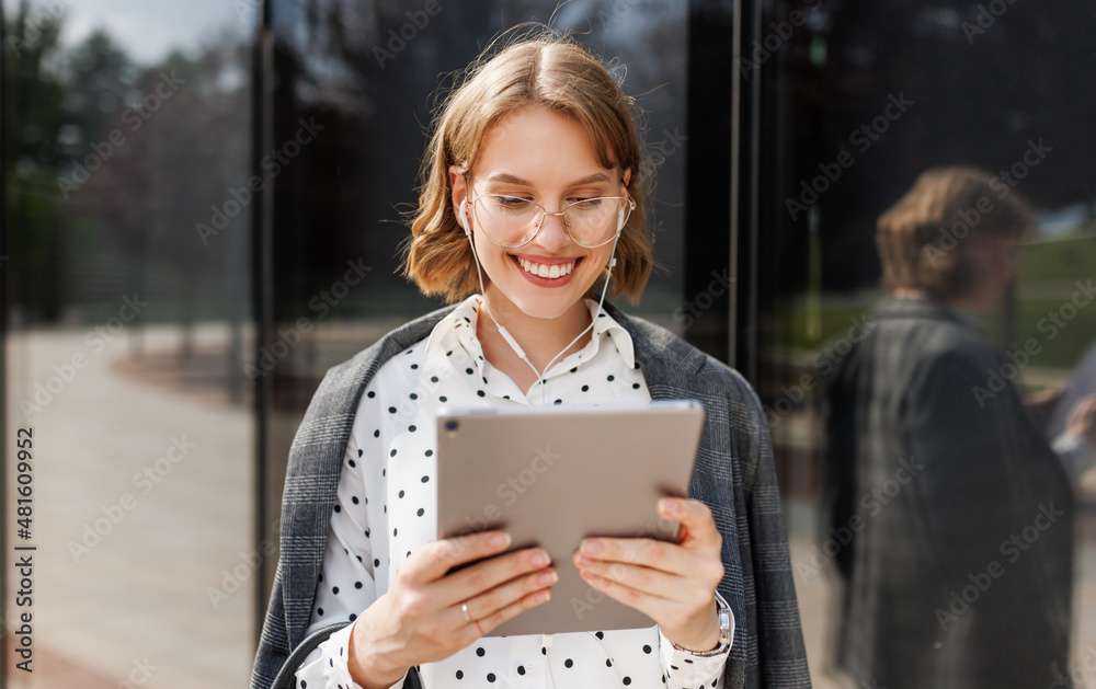 Cheerful business lady in earphones looking at digital tablet while standing on sunny street outdoor