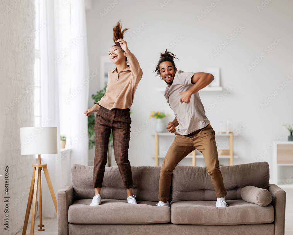 Young overjoyed african american family couple dancing to music on sofa at home