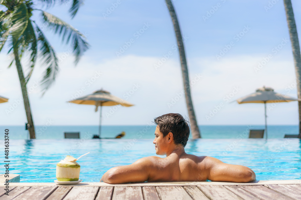 夏季旅游度假概念，旅行者亚洲男子带着椰子在豪华无边泳池酒店放松