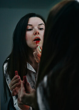 A girl with fair skin paints her lips with red lipstick in the mirror, dressed in a white blouse and an even skirt.