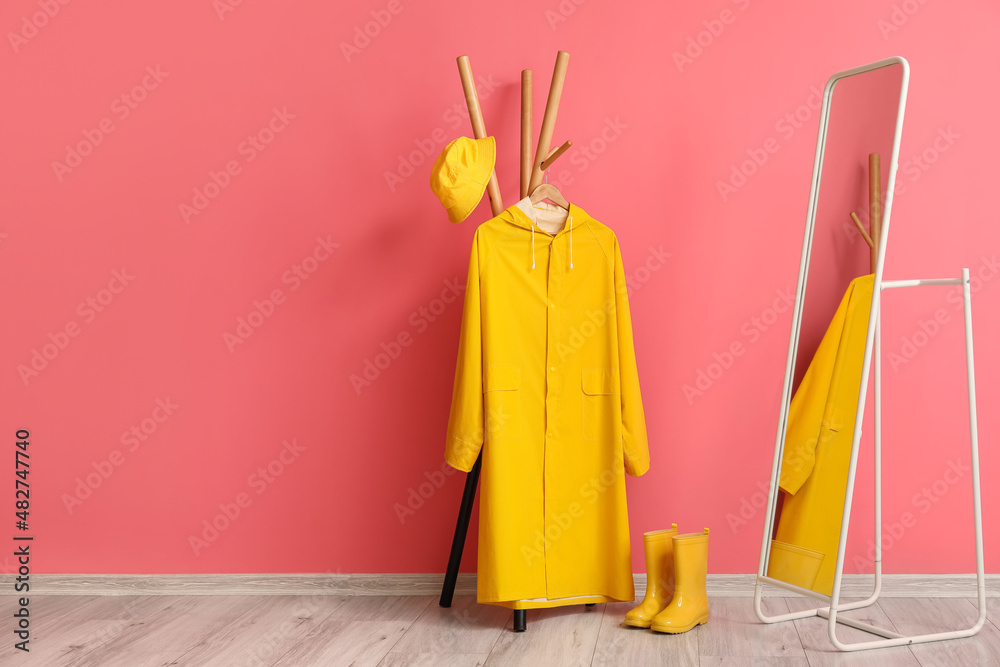 Rack with yellow raincoat, hat, gumboots and mirror near pink wall