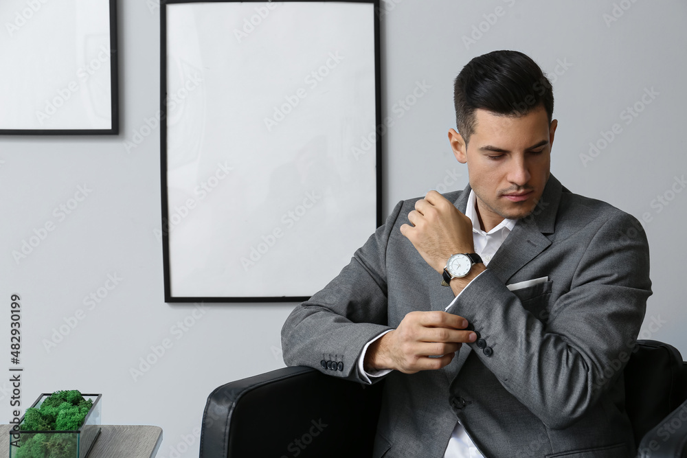 Handsome man in elegant suit sitting in armchair and adjusting sleeve