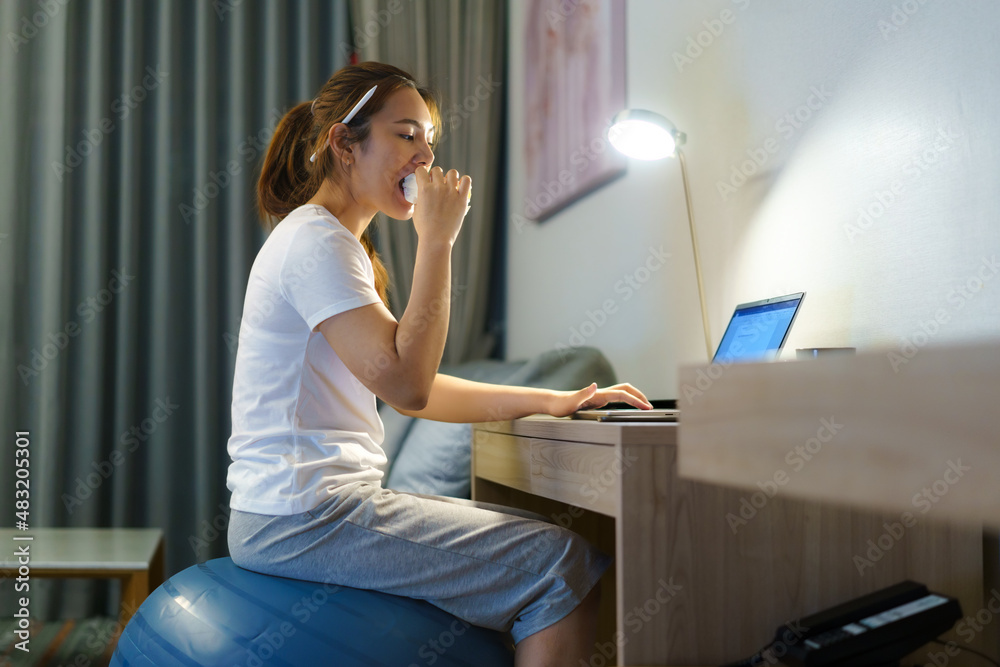 Asian woman relaxing on yoga ball and eating sandwich and chatting online with team of colleagues on