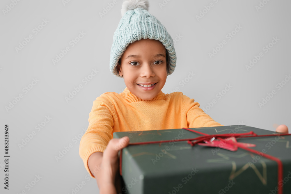 Cute little African-American boy in winter clothes and with Christmas gift on grey background