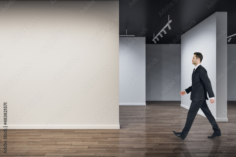 Businessman walking in modern concrete exhibition hall interior with mock up place on wall and woode