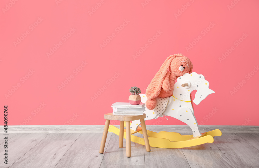 Books, houseplant on table and cute toy rabbit sitting on rocking horse near color wall