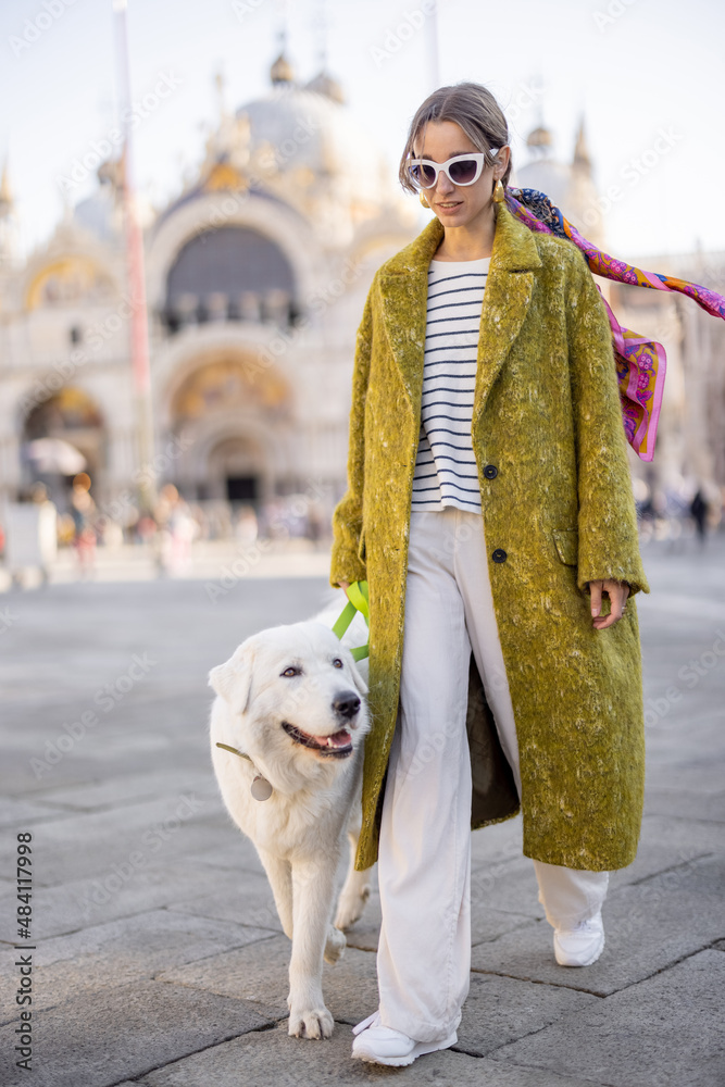 威尼斯年轻时尚女性与白色巨犬的生活方式肖像。穿着外套、戴眼镜的女性