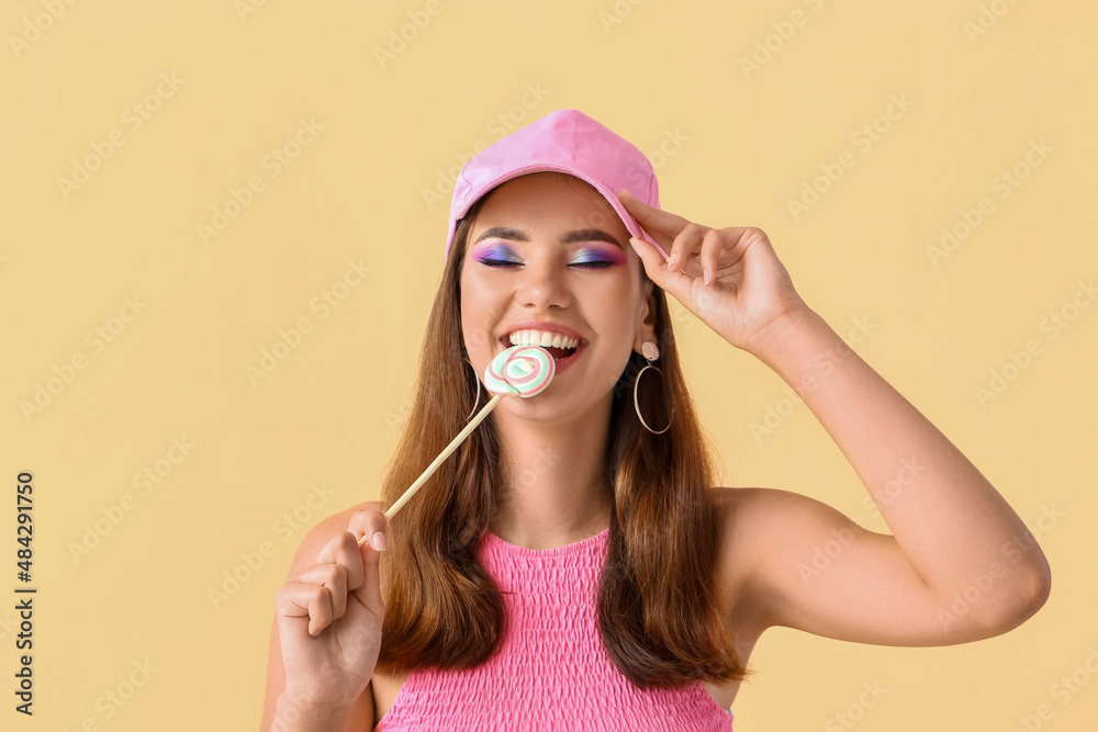 戴粉色帽子的漂亮年轻女子在米色背景下吃甜棒棒糖