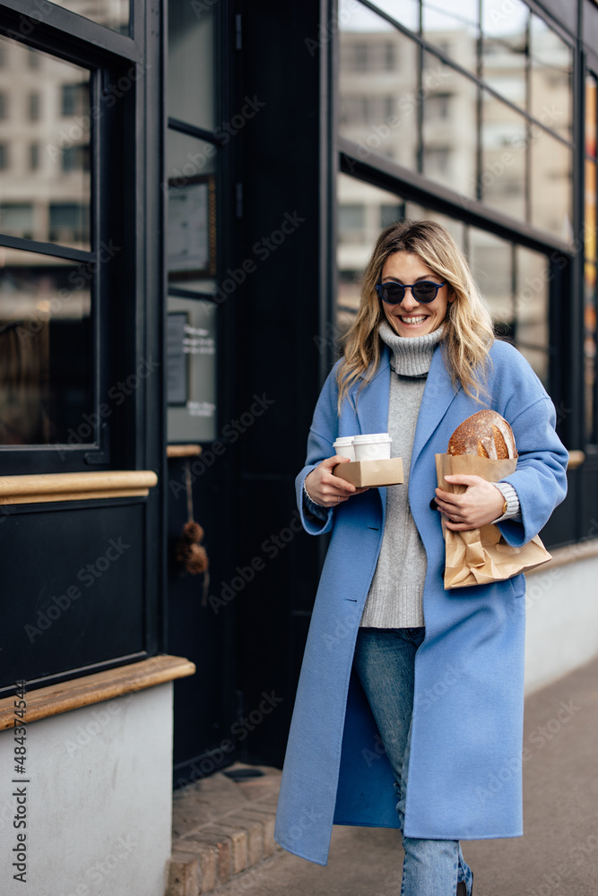 Women having a busy morning, holding cups of coffee and a loaf of bread, walking.