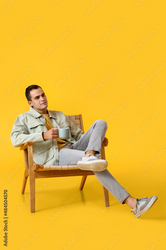 年轻人坐在黄色背景的木制扶手椅上，拿着一杯热饮料休息