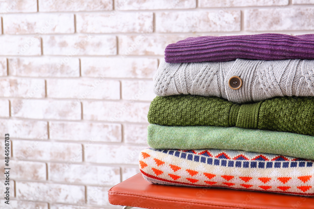 Stack of stylish sweaters on chair against light brick wall