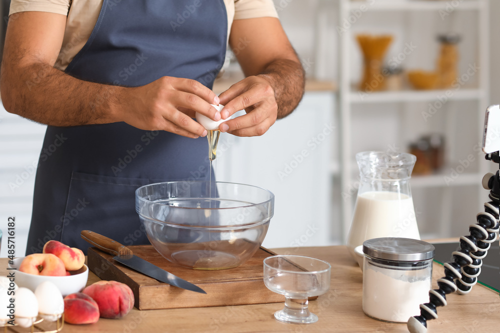 一名年轻男子在厨房里观看烹饪视频教程时将鸡蛋打入碗中，特写镜头