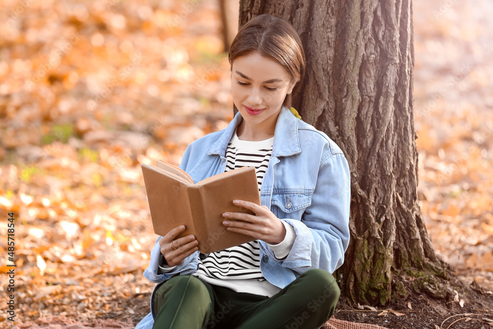 美丽的女人在秋天公园的树旁看书