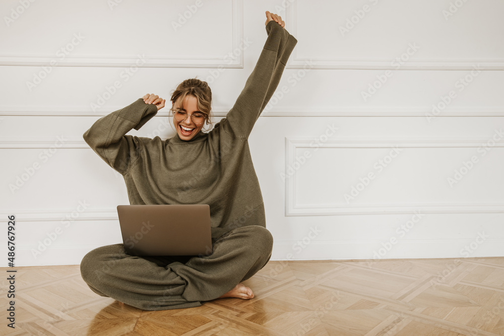 微笑的年轻高加索妇女坐在家里的地板上用笔记本电脑。戴着玻璃的漂亮女孩