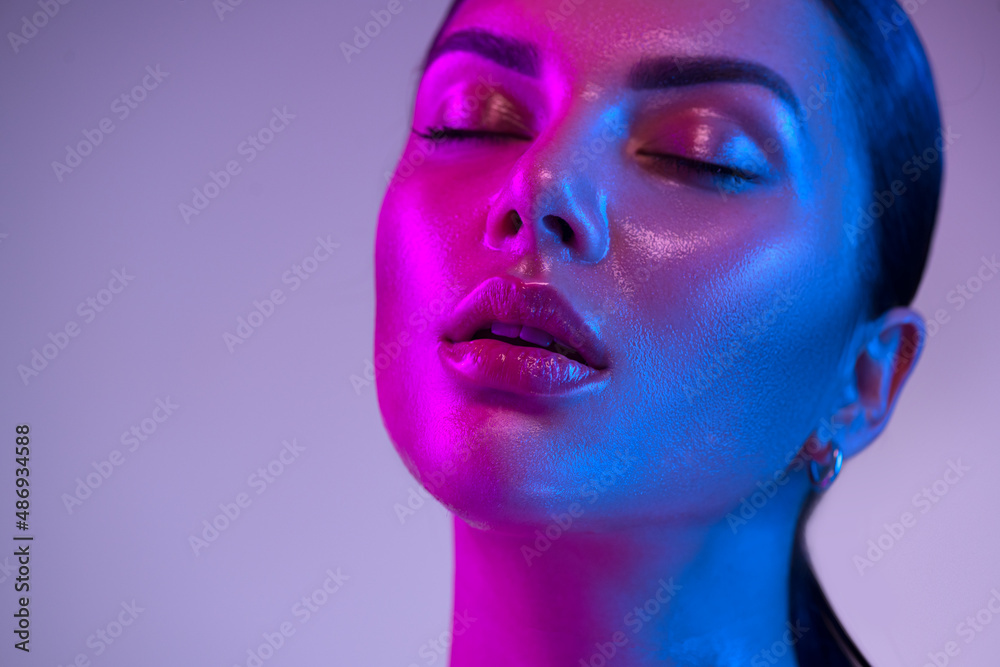 Glossy, wet skin makeup. Fashion model woman face in bright neon colourful lights, beautiful woman, 
