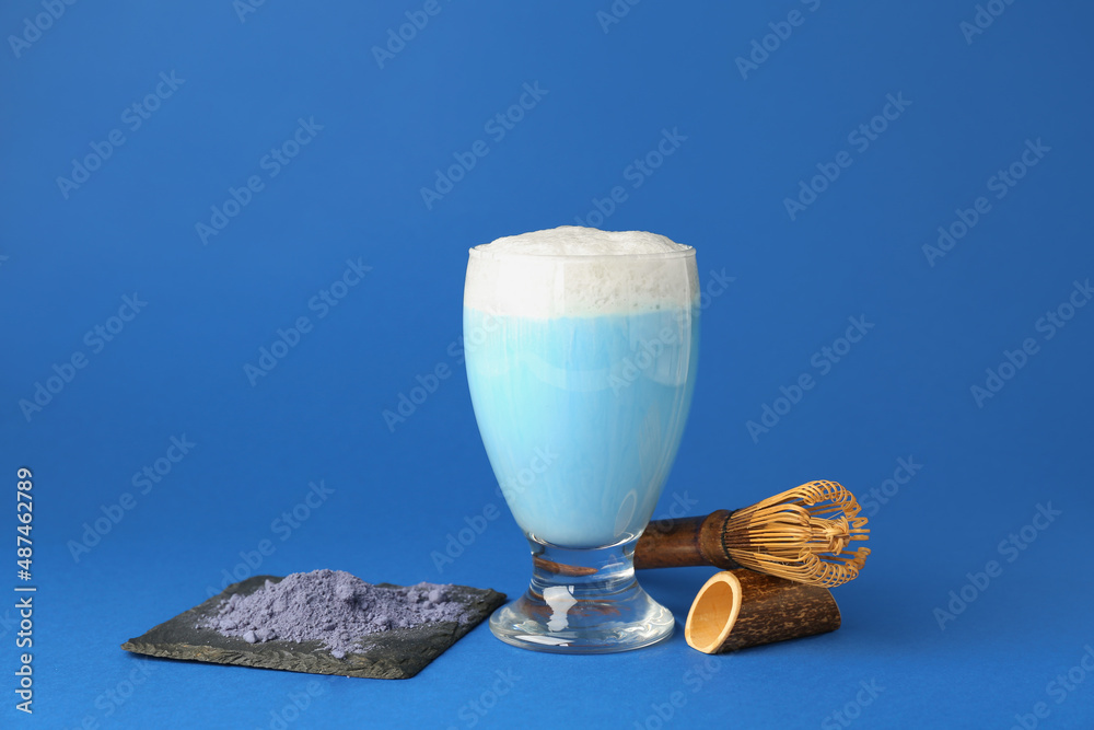 Glass of tasty blue matcha latte, chasen and powder on color background