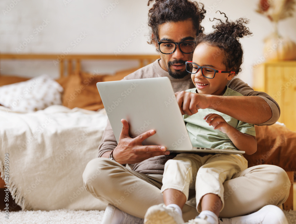 快乐的美国黑人家庭小儿子和爸爸一起在家里用笔记本电脑看有趣的视频