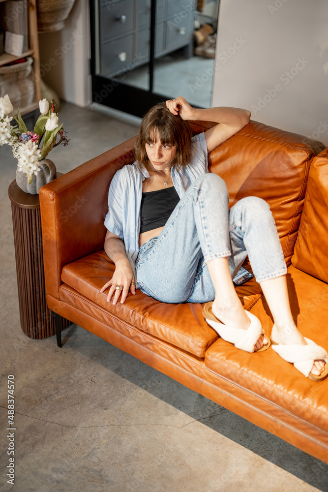 年轻自信的女性在现代家中放松地坐在皮沙发上的生活方式画像。成年人
